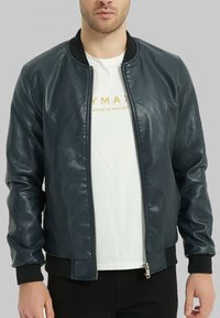 Veste bomber en cuir bleu foncé avec fermeture éclair, poignets et col côtelés noirs. Portée par-dessus un t-shirt blanc avec du texte.