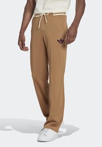 Bruna joggers med en texturerad yta, med en vit snörning och en lila Adidas-logga på vänster lår. Bärs med vita sneakers.