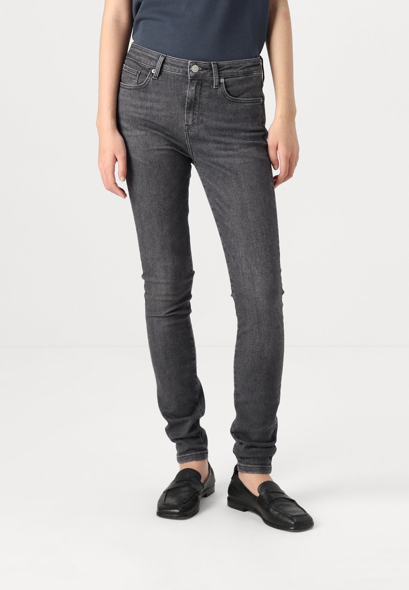 Jeans skinny de color gris oscuro con una cintura de altura media, que cuentan con cinco bolsillos y un cierre de botón. La tela presenta una sutil textura. Zapatos negros sin cordones.