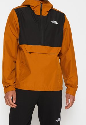 Mężczyzna w pomarańczowo-czarnej kurtce zapinanej do połowy na zamek błyskawiczny oraz czarnych spodniach, obu oznaczonych logo The North Face, stoi na tle jednolitego tła.