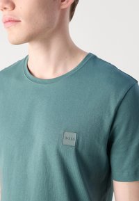 Mėlynas medvilninis marškinėliai su apvaliu kaklu, trumpomis rankovėmis ir kvadratiniu logotipu, turinčiu įstrižas juosteles kairiajame krūtinės kampe.