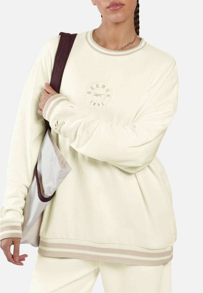 Persona con sudadera y pantalones crema de Reebok, llevando un bolso tote bicolor beige y marrón sobre un hombro, de pie frente a un fondo liso.