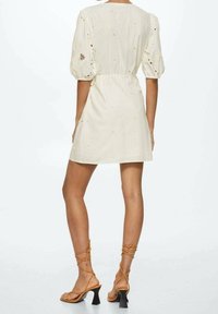 Weißes Kleid aus strukturiertem Stoff, mit Puffärmeln und floraler Stickerei. Getragen mit hellbraunen Riemchensandalen und einem ausgestellten Absatz.
