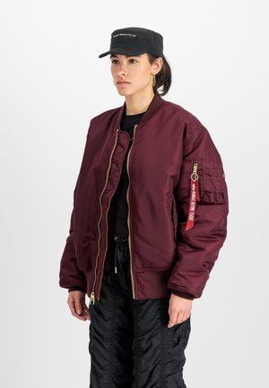 Maroon bomber jacket with gold zipper, featuring side pockets and a red tag on the sleeve. Worn over a black top and paired with black trousers.