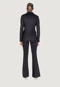 Donna in piedi di spalle, che indossa un blazer nero aderente e pantaloni neri svasati con tacchi alti nudi, su uno sfondo semplice.