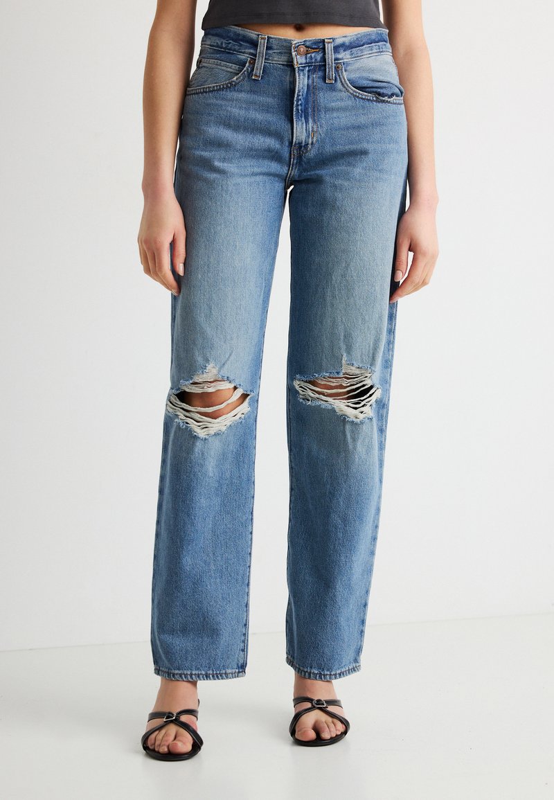 Γυναίκα που φορά μπλε τζιν ψηλόμεσο με μεγάλα σκισίματα και στα δύο γόνατα και μαύρα σανδάλια με τακούνι και ανοιχτά δάχτυλα, στέκεται μπροστά σε λευκό φόντο.