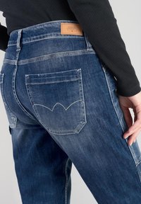 Jeans in denim di un blu scuro con un leggero strappo. Presentano un design cucito della tasca posteriore che ricorda un'onda e un'etichetta in pelle sul girovita.