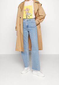 Abrigo trench beige, camiseta gráfica rosa, vaqueros de pernera ancha azul claro y zapatillas blancas. El abrigo cuenta con una cintura ceñida y grandes botones.