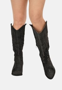 Piernas que llevan botas vaqueras negras hasta la mitad de la pantorrilla, con costuras decorativas y perforaciones, mostradas contra un fondo blanco liso.
