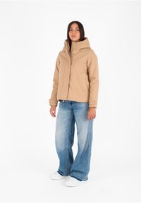 Beige gewatteerde jacket met drukknoopsluiting, zakken aan de voorkant en een gladde textuur, gecombineerd met lichtblauwe wijd-uitlopende jeans en witte sneakers.