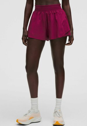Persoon draagt fel magenta sportshorts, witte sportsokken en lichtroze en witte hardloopschoenen, staand tegen een effen lichte achtergrond.