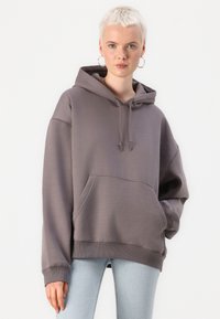 Grå hettegenser med kengurulomme, snorhette og ribbestrikkede cuffs. Overdimensjonert passform med en glatt, myk tekstur. Brukt over lyseblå jeans.