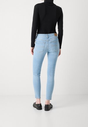 Person trägt hellblaue Skinny-Jeans und ein schwarzes Langarmshirt, steht mit dem Rücken zur Kamera vor einem einfarbigen Hintergrund.