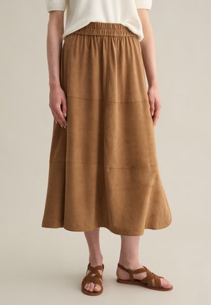 Falda maxi de ante marrón con cintura elástica, con forma acampanada y detalles en las costuras. Combinada con sandalias marrones de tiras.