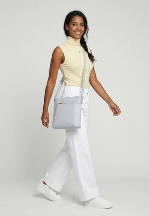 Lächelnde Frau läuft, trägt ein ärmelloses beiges Oberteil, weiße weite Hosen, weiße Sneakers und eine hellgraue Umhängetasche.