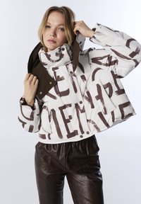 Femme ajustant la capuche d'une doudoune blanche avec de grandes lettres abstraites marron, accompagnée de pantalons texturés marron foncé, sur fond uni.