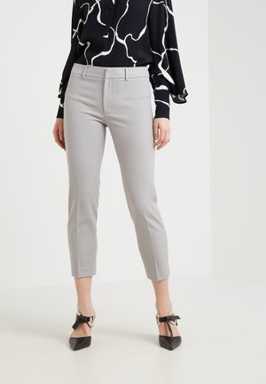 Mujer con pantalones ajustados cortos de color gris claro, blusa negra con estampado y tacones negros de punta con grandes hebillas plateadas y lazos.