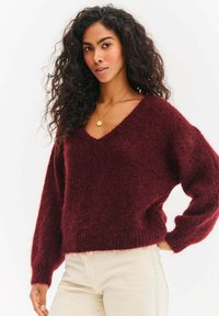 Bordowy, puszysty sweter z dekoltem w serek, szerokimi rękawami i ściągaczem u dołu. Nosi się go na jasne, dopasowane spodnie. Prosty, teksturowany wzór.