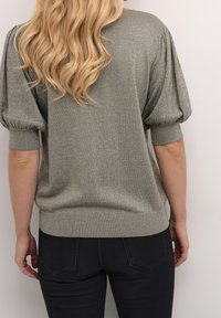 Pull métallisé argenté à manches courtes bouffantes, poignets et ourlet côtelés, associé à un jean foncé. Texture douce, coupe décontractée et finition brillante.