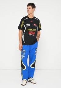 Melnas sporta T-krekls ar logo izdrukām; zilas sporta bikses ar sānu akcentiem un numuriem; valkātas ar baltām un brūnām kedām.