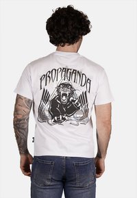 T-shirt bianca in cotone con una grafica nera di un pantera ruggente e il testo "PROPAGANDA" in carattere grassetto sul retro. Maniche corte, vestibilità casual.