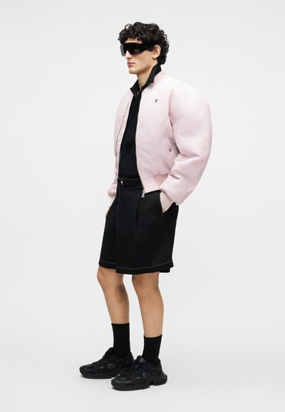 Άνδρας που φοράει υπερμέγεθη μπουφάν bomber σε απαλό ροζ χρώμα, μαύρο πουκάμισο, μαύρες βερμούδες, μαύρα αθλητικά παπούτσια, κάλτσες και μεγάλα μαύρα γυαλιά ηλίου, στέκεται πλαγίως.