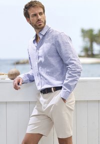 Hellblau gestreiftes Hemd mit Knöpfen, beiges Shirt und ein dunkler Gürtel. Die Umgebung zeigt eine Küstenlandschaft mit Bäumen und einem weißen Geländer.