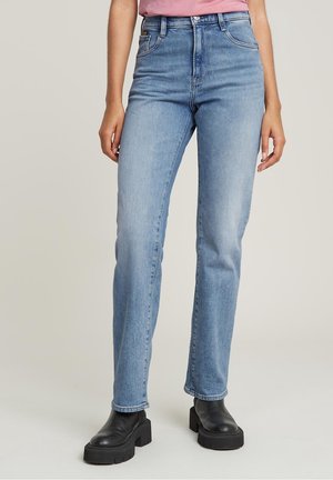 Personne portant un jean bleu clair taille haute à jambes droites et des bottines plateformes noires épaisses, debout devant un fond uni.