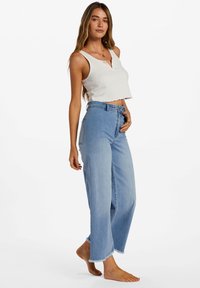 White ribbed crop top with a frayed hem paired with light blue wide-leg jeans featuring raw hem and front pockets. Model stands barefoot.