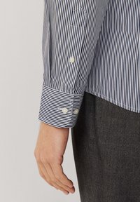 Raidallinen nappikauluspaida, jossa on sinisiä ja valkoisia pystyraitoja, kahden napin hihansuut ja yksi rintatasku. Kangas näyttää sileältä.