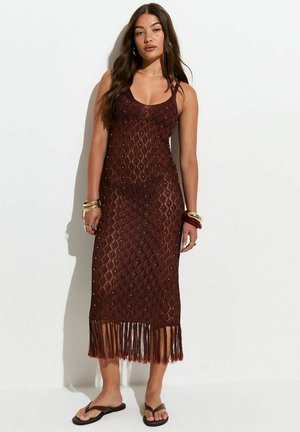 Femme aux longs cheveux portant une robe sans manches en crochet marron foncé avec un ourlet à franges, des bracelets et des tongs foncées, debout contre un mur blanc.
