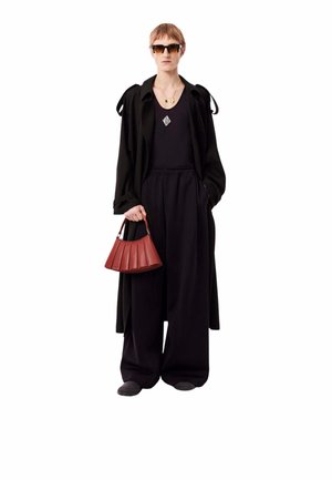 Personne portant un pantalon noir à jambes larges, un haut noir sans manches, un long manteau noir, des lunettes de soleil, et tenant un sac à main rouge plissé.