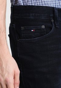 Dunkelblaue Jeans mit schmaler Passform, Fronttaschen und einem kleinen Logo-Patch in Rot, Weiß und Blau auf der Gesäßtasche. Strukturiertes Gewebe.