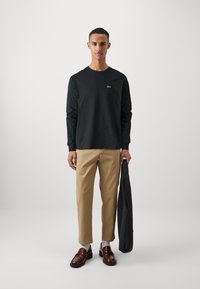 Black long-sleeve shirt, khaki trousers, and brown loafers. Shirt features a small green logo. Model holds a dark jacket. Neutral background.