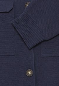 Pull bleu marine avec un tricot texturé. Il comporte deux poches avant et des poignets côtelés. Comprend des boutons marron pour la fermeture.