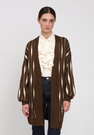 Frau trägt eine braune gemusterte lange Strickjacke über einer weißen Rüschenbluse und dunkelblauer Hose, steht vor einfarbig hellem Hintergrund.