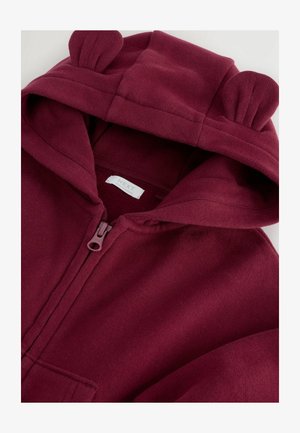 Burgundi színű, cipzáras kapucnis pulóver, amely bolyhos anyagból készült, kapucnija medvefülekkel díszített, és egy kis cimke található benne. Puha tapintású, egyszerű dizájnnal rendelkezik.