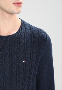Nahaufnahme einer Person, die einen dunkelblauen, strukturierten Pullover mit einem kleinen rot-weiß-blauen Logo auf der Brust trägt, vor einem einfarbigen Hintergrund.