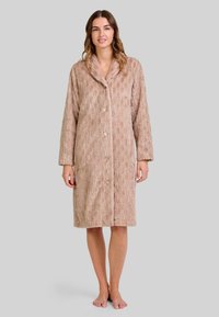 Mujer de pie descalza, vistiendo una bata de botones de largo hasta la rodilla, texturizada, en color beige, con mangas largas y cuello de chal.