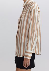 Camicia a maniche lunghe a righe in beige e bianco, realizzata in tessuto liscio. Presenta un polsino con bottoni e un colletto classico. Modello con gonna nera.