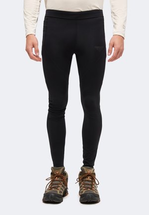 Uomo con leggings neri aderenti e scarponi da trekking marroni, in piedi con le mani rilassate lungo i fianchi.
