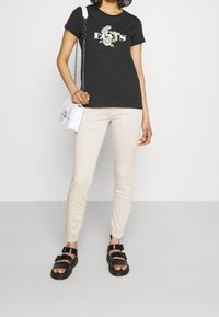 Czarna koszulka z kwiecistym wzorem, beżowe dopasowane spodnie z kieszeniami i czarne sandały. Mała biała torba na ramię z łańcuszkowym paskiem.