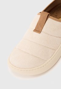 Beige slip-on cipő párnázott textúrájú felsőrésszel, barna szövet húzófüllel és krémszínű gumitalppal fehér háttéren.