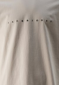 Camiseta gris claro hecha de una tela suave, con un pequeño logo impreso en negro "JACK&JONES" centrado en el frente. Diseño sencillo de cuello redondo.