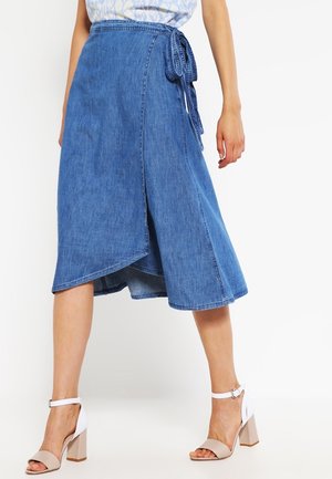 Femme portant une jupe portefeuille en jean bleu nouée à la taille, associée à des sandales à talons hauts beige et blanches, debout sur un fond blanc.