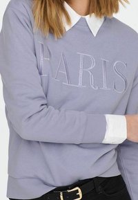 Felpa di un leggero color viola con la scritta ricamata "PARIS", dotata di scollatura tonda e polsini a costine, indossata sopra una camicia bianca con colletto.