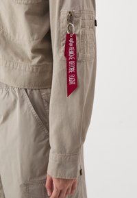 Hellbeige Jacke mit einer Reißverschlusstasche und rotem Stoffetikett mit der Aufschrift "REMOVE BEFORE FLIGHT." Glatte Textur, funktionales Design.