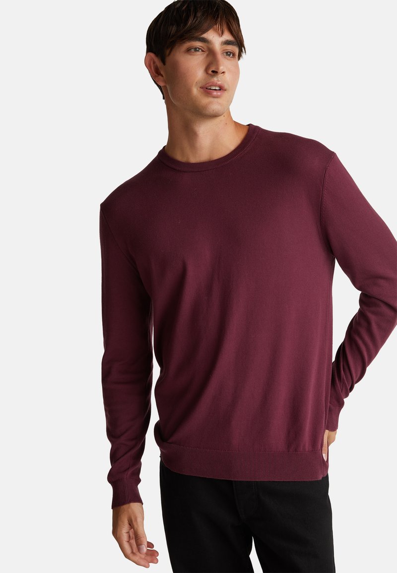 Bordeaux langetermijntrui gemaakt van zacht breimateriaal, met een ronde hals, geribde manchetten en een slim-fit ontwerp tegen een effen achtergrond.