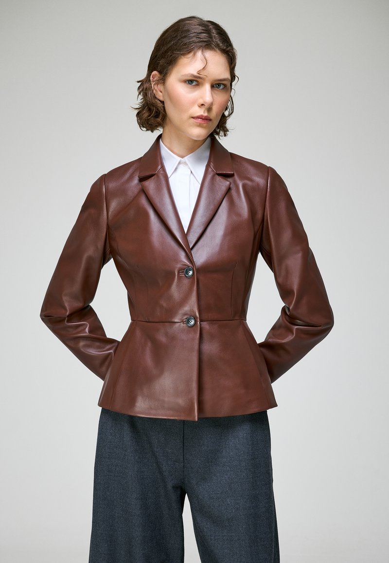 Jeune personne aux cheveux courts portant un blazer en cuir marron sur mesure par-dessus une chemise blanche et un pantalon gris à jambes larges, debout les mains derrière le dos.