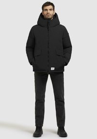 Sort polstret jakke med høj krave, lynlåslukning og to forlommer. Båret over sorte jeans og sorte støvler. Simpelt, minimalistisk design.
